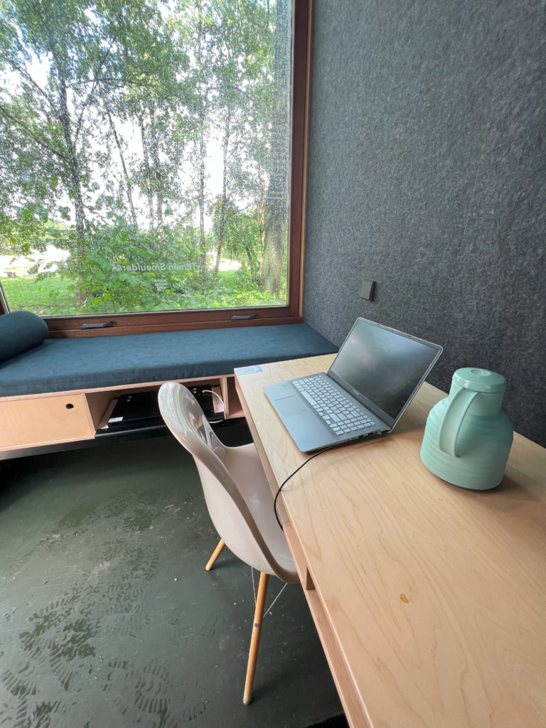 Een foto van de inrichting van de tiny office. Je ziet een zitvensterbank bij het raam, een houten burau en een witte stoel. Op het bureau staat een laptop en een thermoskan.